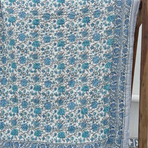May include: A white fabric with a blue floral pattern and a blue border. The floral design features various shades of blue and teal flowers with grey stems and leaves. The fabric is draped over a wooden frame.