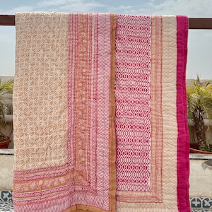 May include: A pink and beige cotton quilt with a geometric pattern. The quilt is folded and hanging on a wooden railing.