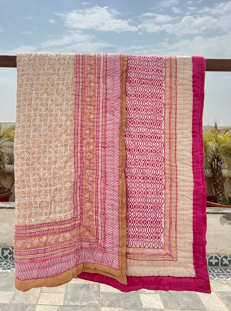 May include: A pink and beige cotton quilt with a geometric pattern. The quilt is folded and hanging on a wooden railing.