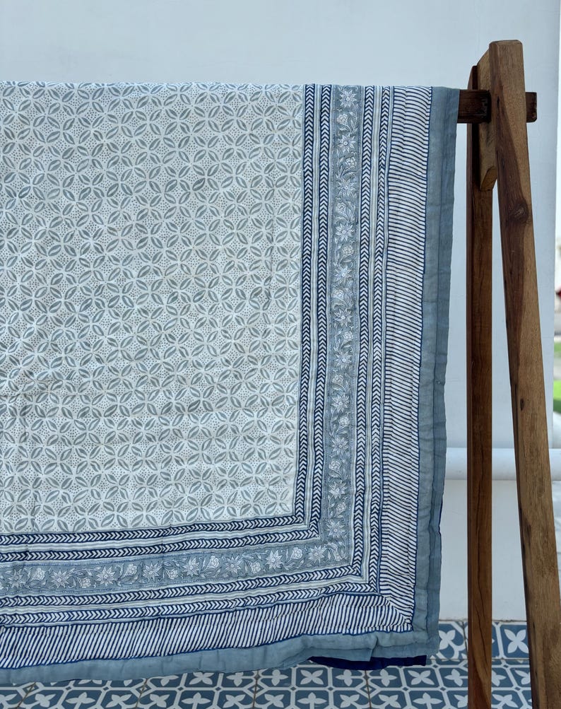 May include: A white and blue cotton quilt with a geometric pattern. The quilt has a border with a floral design. The quilt is folded and draped over a wooden stand.