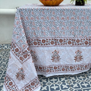 May include: White tablecloth with a floral pattern in shades of brown and red. The tablecloth is draped over a table, with a basket of apples and a small potted plant on top. The tablecloth has a detailed border.