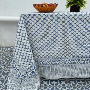 May include: White tablecloth with a blue geometric pattern and floral border. A woven basket of apples and a potted plant sit on the table. The tablecloth has a repeating chain-link design.
