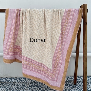 May include: A cream-colored cotton dohar with a geometric pattern and a pink and brown border. The dohar is folded and draped over a wooden rack.