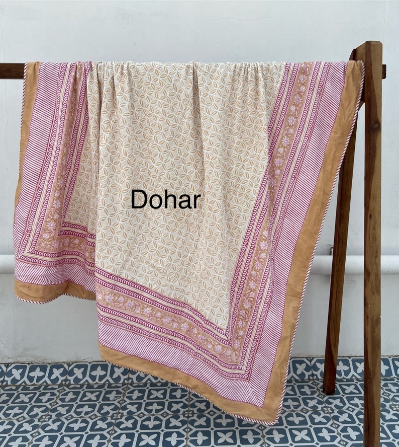 May include: A cream-colored cotton dohar with a geometric pattern and a pink and brown border. The dohar is folded and draped over a wooden rack.