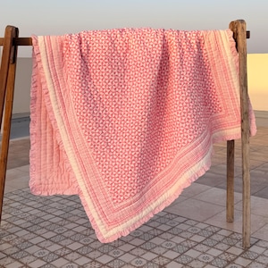 Op de afbeelding: Een roze en wit patroon quilt met een geometrisch ontwerp, weergegeven op een houten droogrek. De quilt heeft een gedetailleerde rand en franjes langs de randen. De quilt is gemaakt van zacht materiaal.