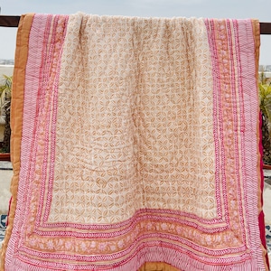 May include: A beige and pink cotton quilt with a floral pattern. The quilt has a border with a repeating pattern of pink and white stripes and a floral design.