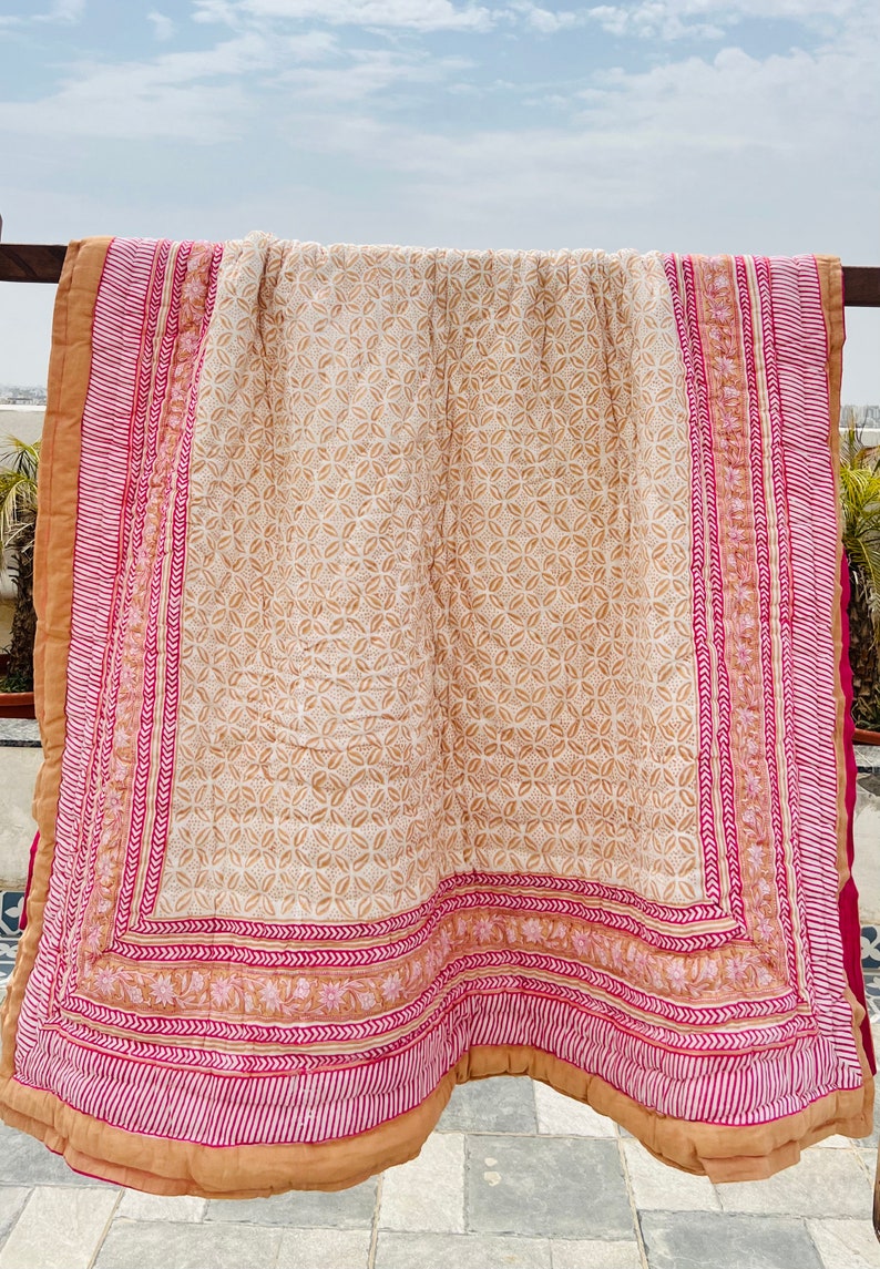 May include: A beige and pink cotton quilt with a floral pattern. The quilt has a border with a repeating pattern of pink and white stripes and a floral design.