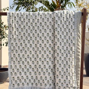 May include: A white quilt with a repeating floral pattern in olive green and gray. The quilt has a striped border with a ruffled edge. The quilt is displayed on a wooden rack outdoors.
