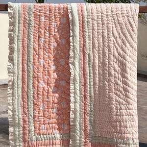 May include: A quilted blanket with a floral pattern in shades of peach and white, accented by vertical stripes of light green and cream. The edges are finished with a ruffled trim, creating a soft and inviting aesthetic. The blanket is displayed outdoors.