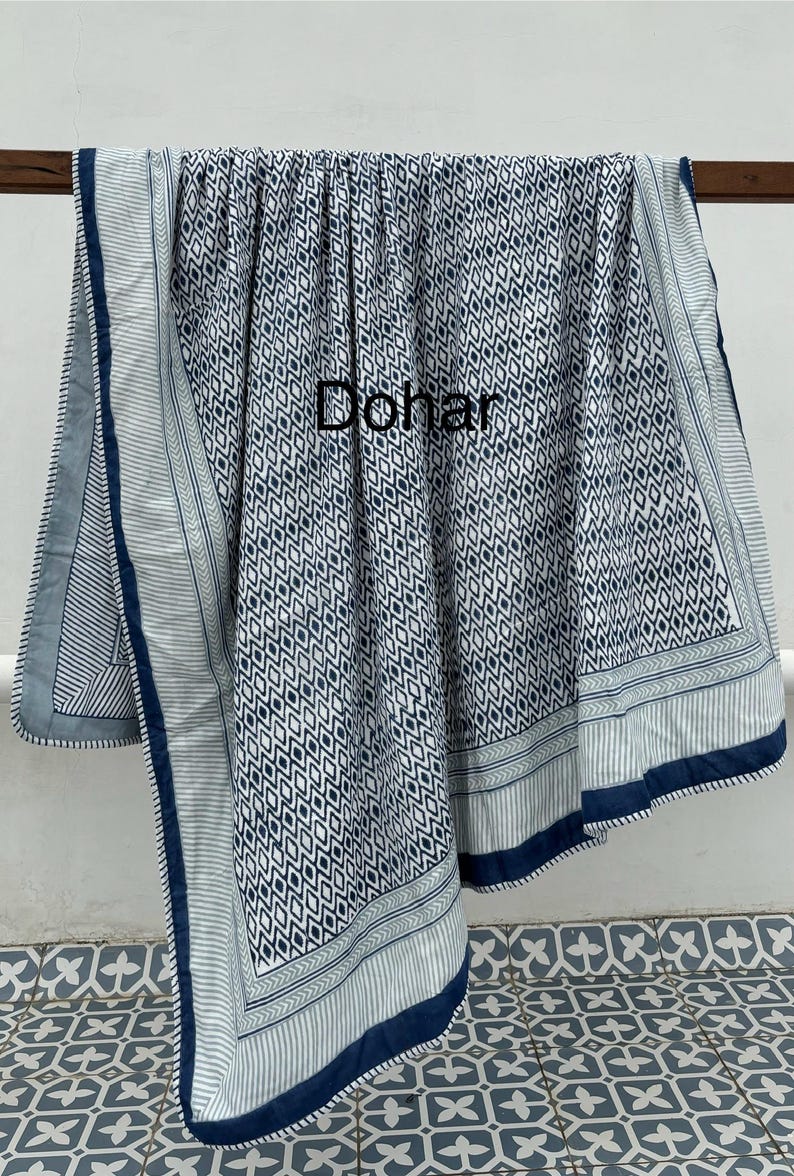 May include: A blue and white patterned cotton quilt with a geometric design. The quilt has a blue border with a white stripe and a green stripe. The word "Dohar" is written in black on the quilt.