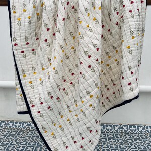 May include: A white quilted blanket with a floral pattern of red and yellow flowers and green leaves. The blanket has a navy blue border and is draped over a wooden rod. The quilt is approximately 70 inches long and 60 inches wide.