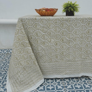 May include: A rectangular tablecloth with a repeating olive green and white floral pattern. The tablecloth is draped over a table, with a woven basket of apples and a small potted plant on top. The tablecloth has a white border.