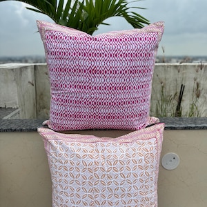 May include: Two square throw pillows with a white background and colorful geometric patterns. The top pillow has a pink diamond pattern and the bottom pillow has a light brown floral pattern.