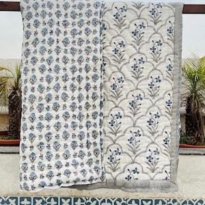 May include: Two white quilts with blue floral patterns. One quilt has a repeating pattern of small flowers, while the other features a scalloped design with larger floral motifs. Both have gray borders.