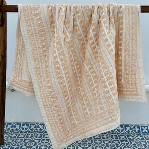 May include: A cream-colored quilt with a repeating pattern of vertical stripes and floral motifs in a warm, light brown hue. The quilt is draped over a wooden rack, showcasing its detailed design and texture. The edges have a decorative border.