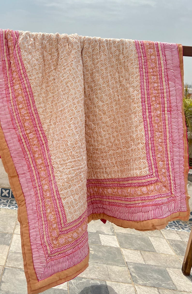 May include: A beige and pink cotton quilt with a floral pattern. The quilt has a border with a repeating pattern of pink and white stripes.