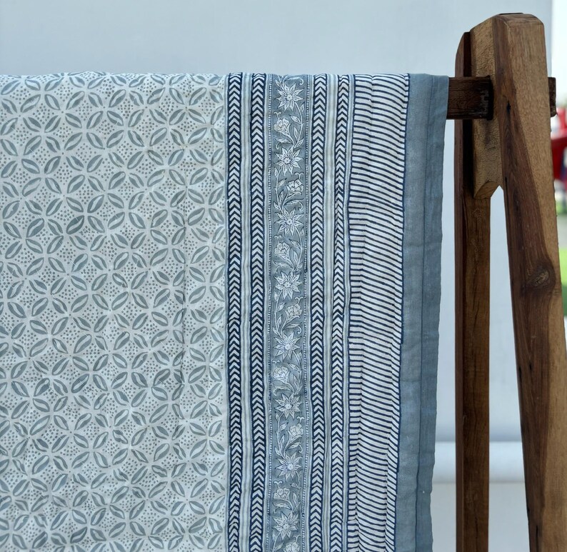 May include: A blue and white cotton quilt with a floral and geometric pattern. The quilt is hanging on a wooden rack.