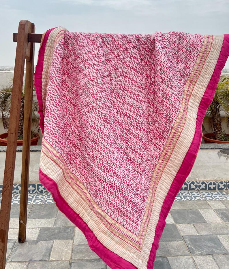 May include: A pink and white kantha quilt with a geometric pattern. The quilt has a cream border with gold embroidery and a pink border.