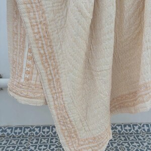 May include: Cream-colored quilted blanket with a repeating floral pattern. The blanket has a border with a geometric design in a light brown hue. The blanket is draped over a surface, showcasing its texture and design.