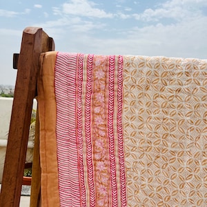 May include: A pink and brown patterned cotton quilt with a white background. The quilt is folded and draped over a wooden chair.