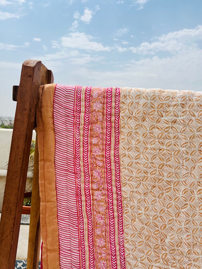 May include: A pink and brown patterned cotton quilt with a white background. The quilt is folded and draped over a wooden chair.