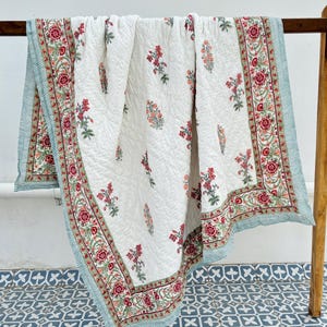Peut inclure: Une courtepointe blanche avec un motif floral rouge, vert et pêche. La courtepointe a une bordure rayée bleu clair et blanche. La courtepointe est exposée sur un support en bois contre un mur blanc.