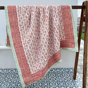 May include: A floral patterned quilt with a white background and red flowers. The quilt has a red border with a green geometric pattern. The quilt is displayed on a wooden rack.
