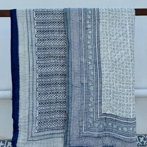 May include: Two blue and white patterned cotton quilts with a geometric design. The quilts are folded and hanging on a wooden rod.
