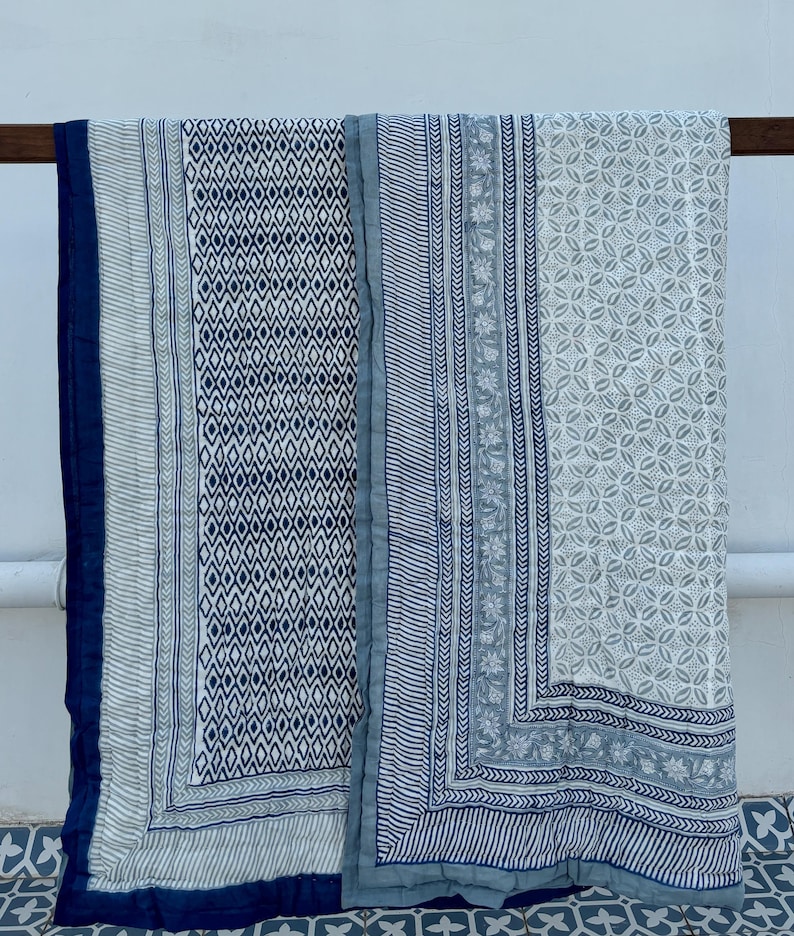 May include: Two blue and white patterned cotton quilts with a geometric design. The quilts are folded and hanging on a wooden rod.