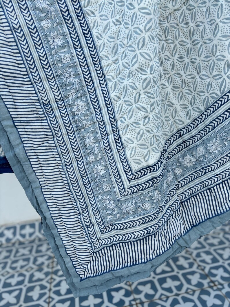 May include: A blue and white cotton quilt with a floral pattern and a geometric border. The quilt is folded in half and the corner is visible.