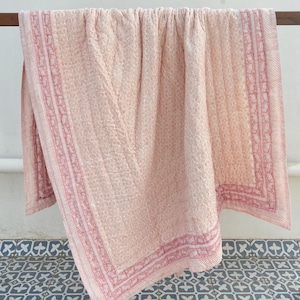 Könnte beinhalten: Eine rosafarbene und weiße Baumwolldecke mit einem Blumenmuster und einem rosafarbenen Rand. Die Decke hängt an einer Holzstange.