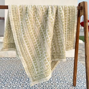 Op de afbeelding: Een lichtgroen en wit patroonquiltdoek gedrapeerd over een houten rek. De quilt heeft verticale strepen met een herhalend bladontwerp. De randen zijn afgewerkt met een rand. De quilt is gemaakt van een zacht, lichtgewicht materiaal.