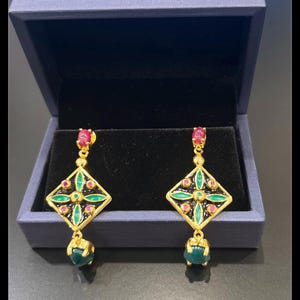 May include: Gold-coloured earrings with a geometric design. Each earring has a red gemstone at the top, a black and green diamond-shaped centre with red and green accents, and a green gemstone at the bottom. The earrings are displayed in a blue jewellery box.