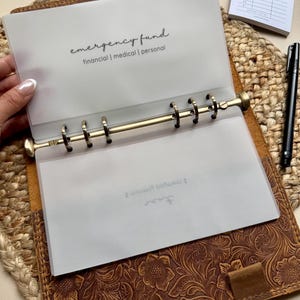 May include: A clear plastic sheet with the text "emergency fund" and "financial | medical | personal" printed on it. The sheet is inserted into a brown leather binder with a floral embossed design.
