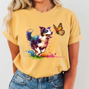 Może przedstawiać: Żółta koszulka z kolorową grafiką przedstawiającą border collie biegnącego w kierunku motyla. Motyl jest pomarańczowy i czarny z żółtym tłem.