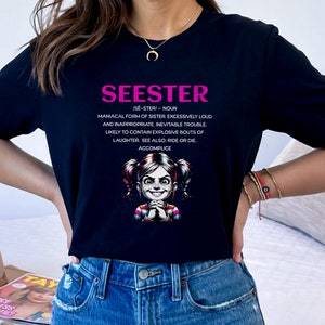 以下が含まれることがあります： 「SEESTER」と書いてあるピンクのグラフィックが施された黒いTシャツ。この言葉は、「妹の狂騒的な形態、過度に騒々しく不適切で、避けられない問題、爆発的な笑い声を起こしやすい。また、生きるか死ぬか。共犯者。」と定義されています。