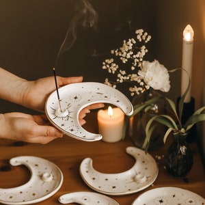 May include: White ceramic moon-shaped incense holders with gold star and sunburst designs. The holders have a small hole for incense sticks. Incense is burning in one holder, with smoke rising. Other holders are arranged on a wooden surface.