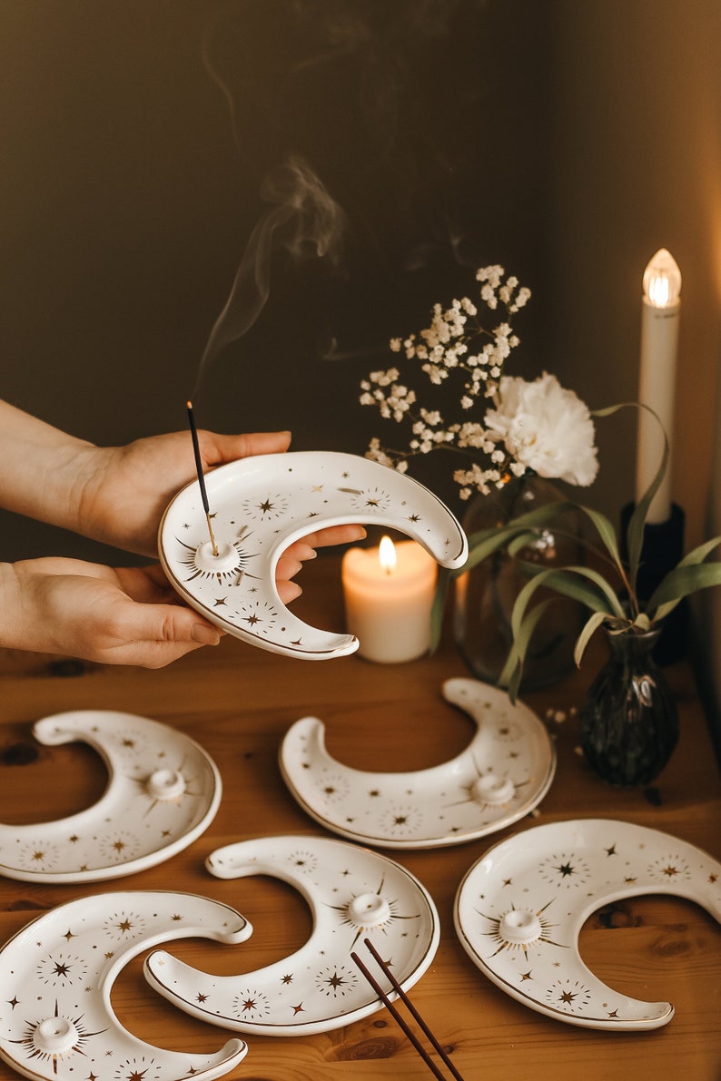 May include: White ceramic moon-shaped incense holders with gold star and sunburst designs. The holders have a small hole for incense sticks. Incense is burning in one holder, with smoke rising. Other holders are arranged on a wooden surface.