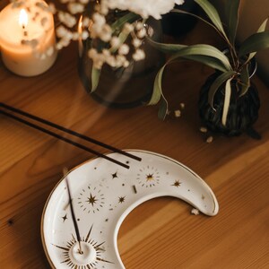 May include: White crescent moon-shaped incense holder with gold star and sunburst designs. Two incense sticks rest on the holder. The holder is on a wooden surface, with a lit candle and floral arrangements in the background.