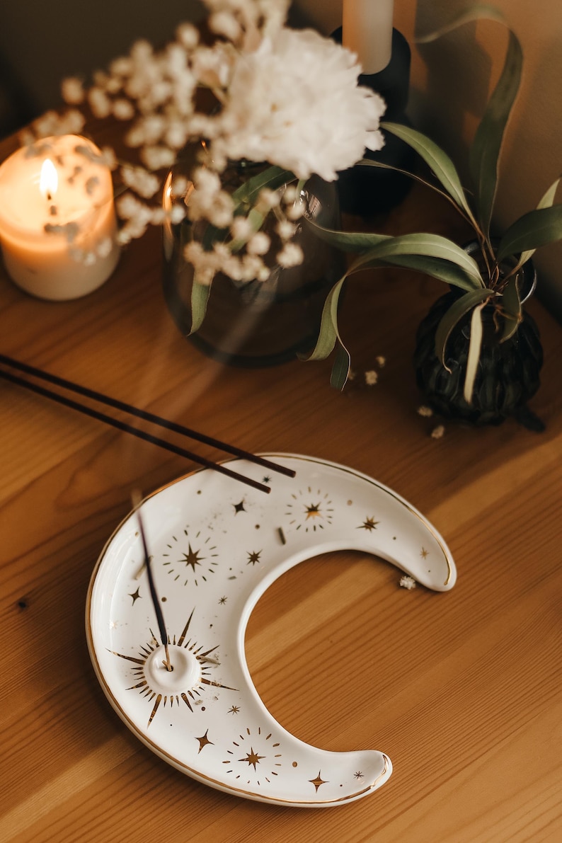 May include: White crescent moon-shaped incense holder with gold star and sunburst designs. Two incense sticks rest on the holder. The holder is on a wooden surface, with a lit candle and floral arrangements in the background.
