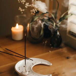 May include: A crescent-shaped, white ceramic incense holder with gold star and sunburst designs. Incense sticks are placed in the holder, with smoke rising. A lit candle and flowers are in the background, creating a relaxing atmosphere.