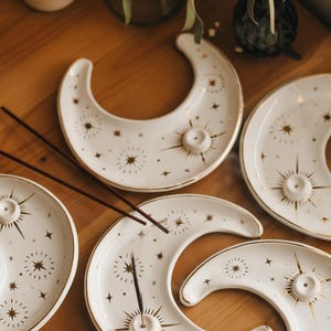 May include: White ceramic moon-shaped incense holders with gold star and sunburst designs. The incense holders have a small hole for incense sticks. The holders are arranged on a wooden surface.