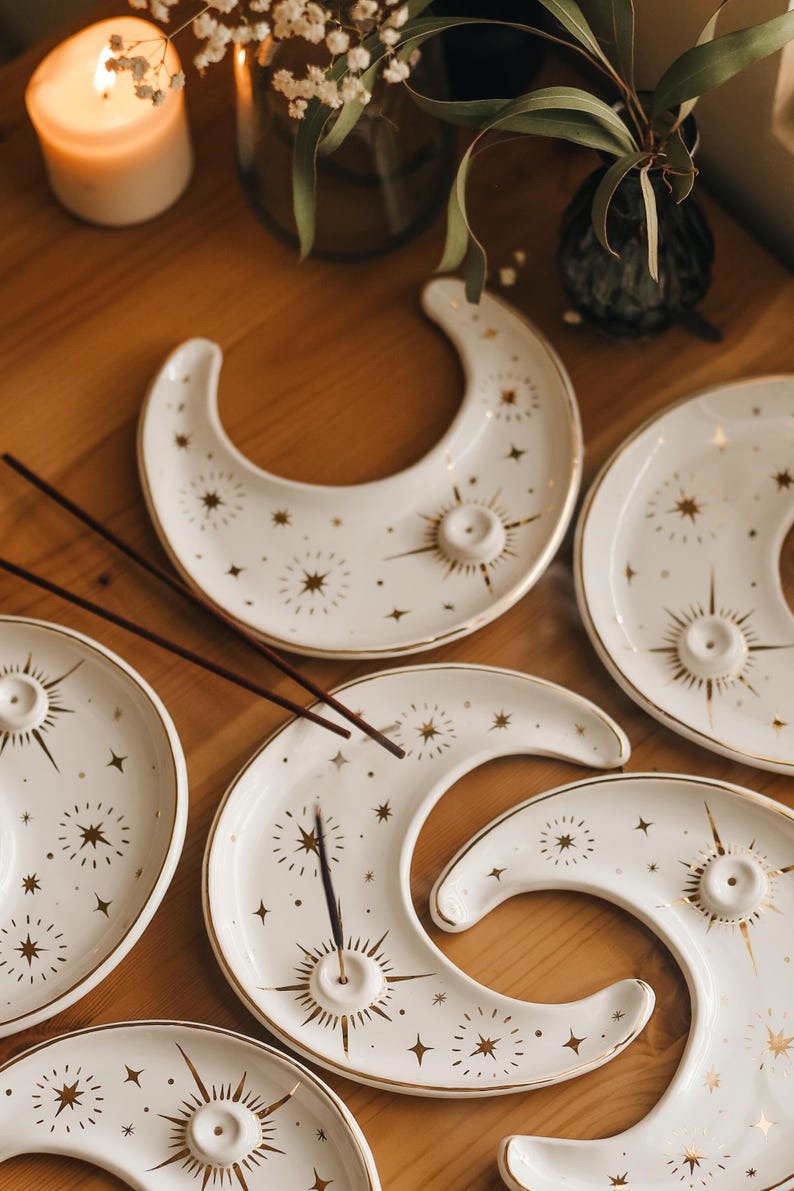 May include: White ceramic moon-shaped incense holders with gold star and sunburst designs. The incense holders have a small hole for incense sticks. The holders are arranged on a wooden surface.