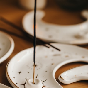 May include: White ceramic incense holder in a crescent moon shape, adorned with gold starburst and star details. An incense stick is lit and placed in the holder. The background includes a lit candle and other holders.