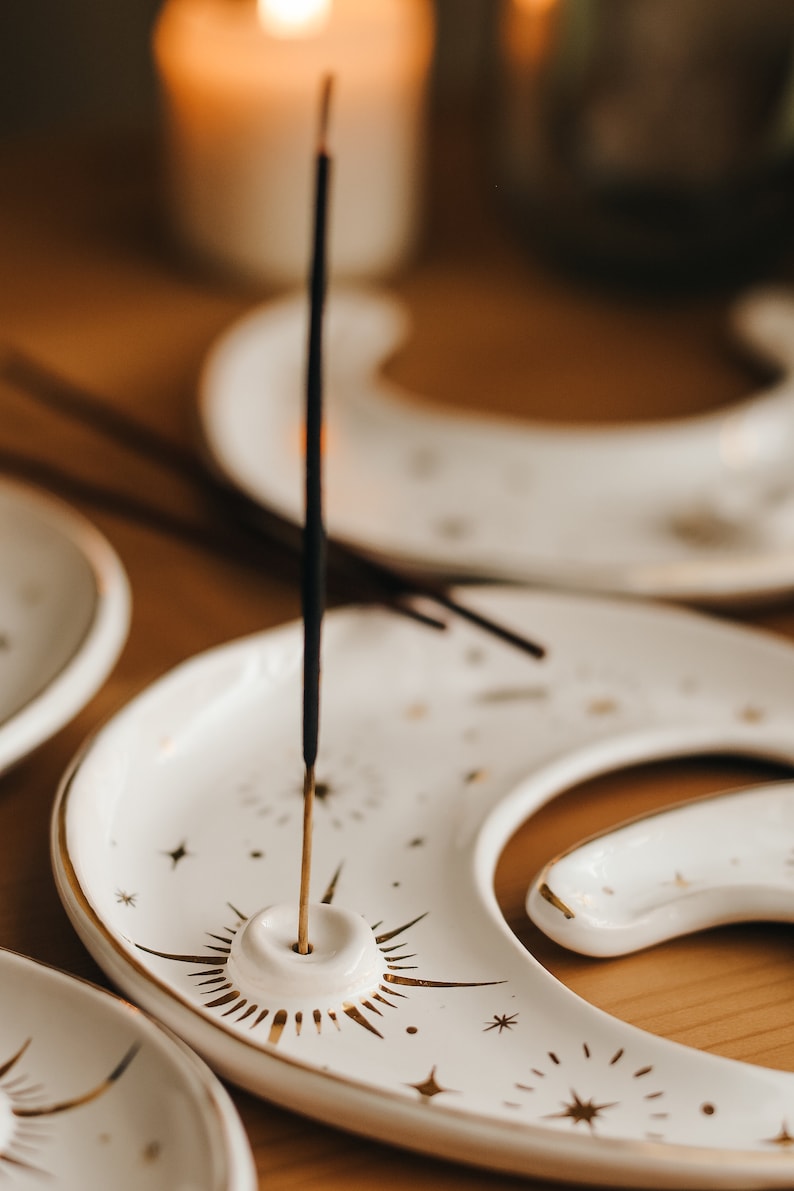 May include: White ceramic incense holder in a crescent moon shape, adorned with gold starburst and star details. An incense stick is lit and placed in the holder. The background includes a lit candle and other holders.