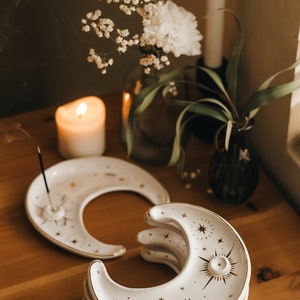May include: A stack of white ceramic moon-shaped incense holders with gold star and sunburst designs. The holders are on a wooden surface, with a lit candle, flowers, and a vase in the background. The incense holder is approximately 5 inches long.