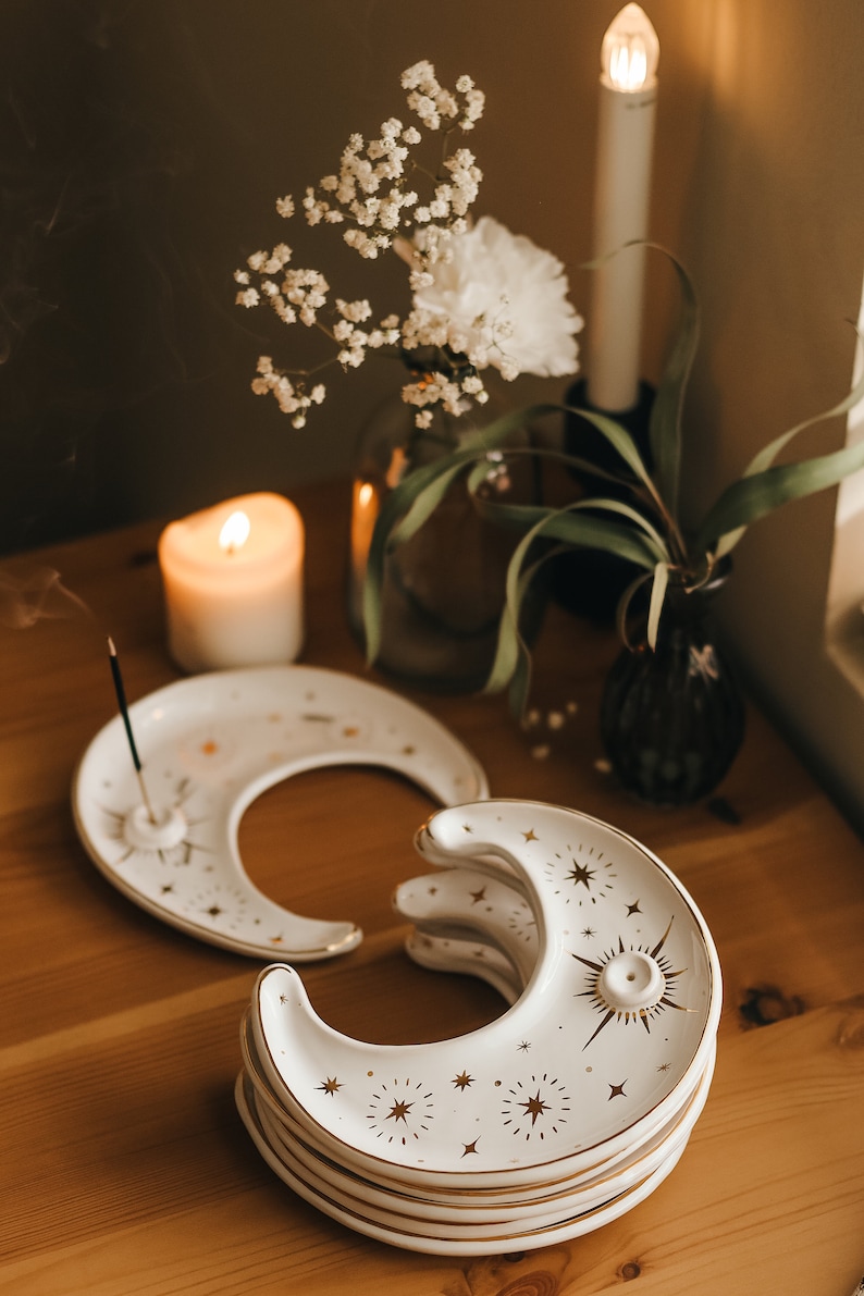 May include: A stack of white ceramic moon-shaped incense holders with gold star and sunburst designs. The holders are on a wooden surface, with a lit candle, flowers, and a vase in the background. The incense holder is approximately 5 inches long.