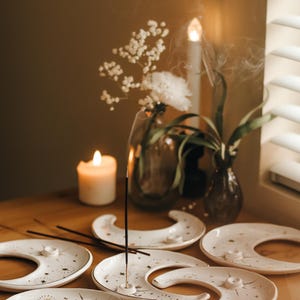May include: A collection of white ceramic moon-shaped incense holders with gold star and line designs. Incense sticks are placed in the holders, with smoke rising. A lit candle and flowers in vases are also on the wooden surface.