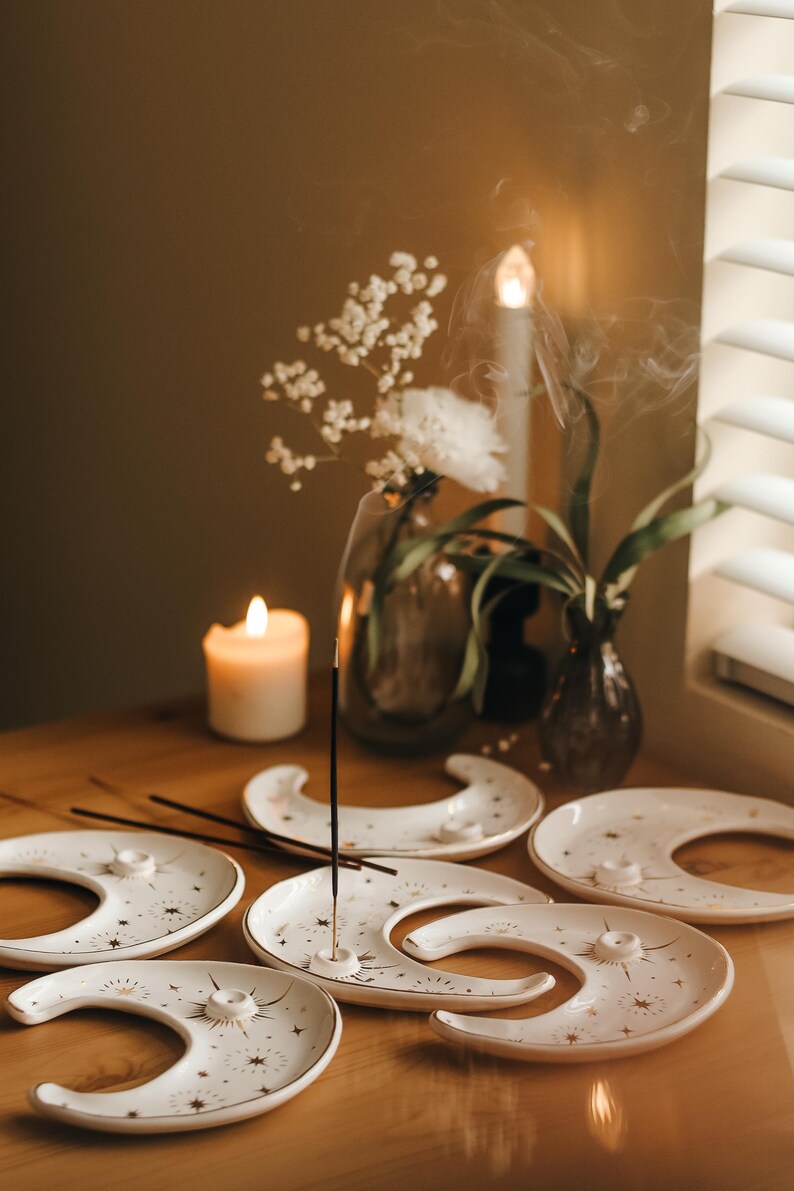 May include: A collection of white ceramic moon-shaped incense holders with gold star and line designs. Incense sticks are placed in the holders, with smoke rising. A lit candle and flowers in vases are also on the wooden surface.