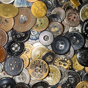 以下が含まれることがあります： 様々なサイズ、形、色のヴィンテージボタンのコレクション。ボタンは金属とプラスチック製で、「SWEET ORR」や「CROWN OVERALL」などのエンボス加工されたテキストが施されています。黒、金、銀、ブロンズの色合いが含まれています。